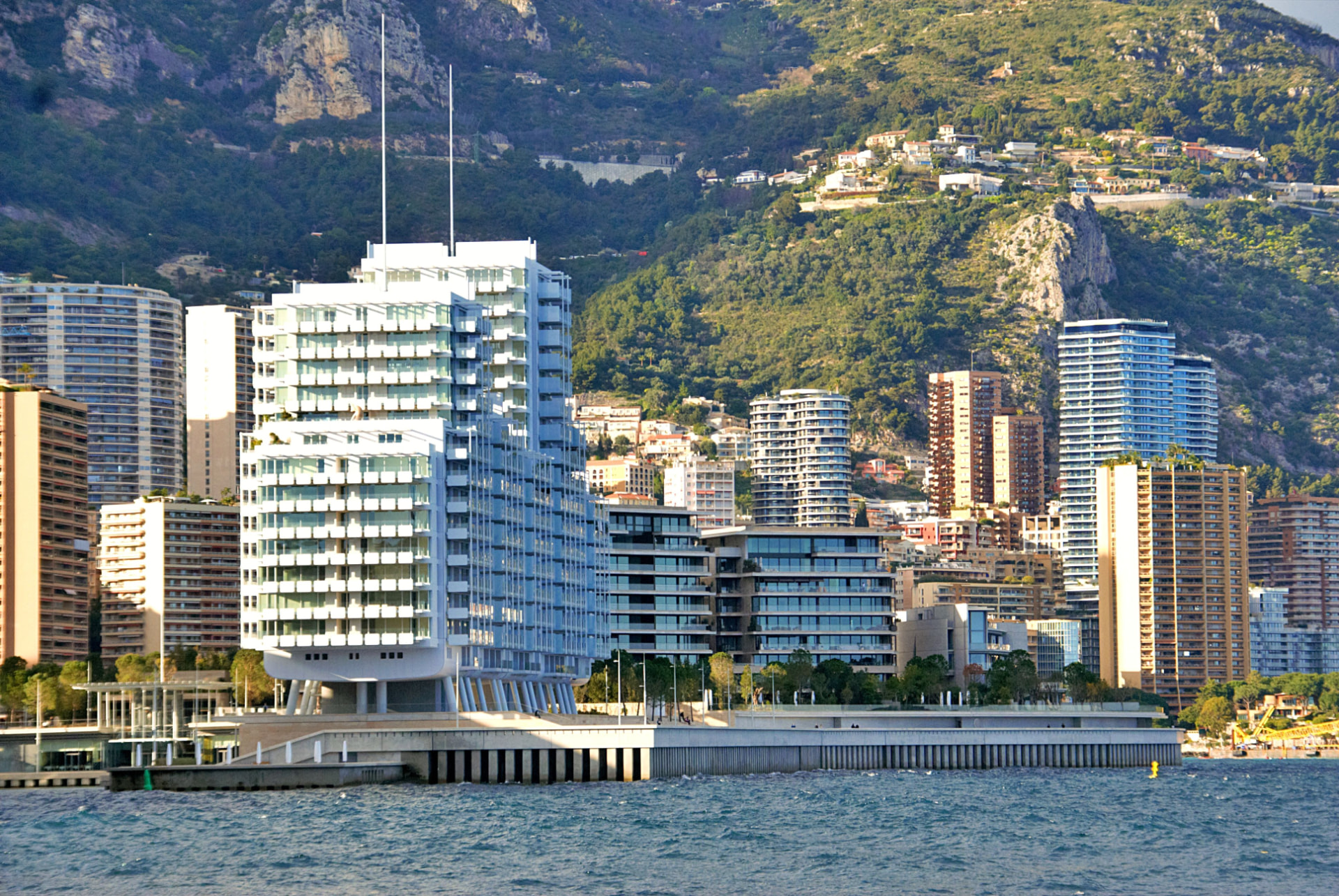 Le Renzo, a multi-tiered white residential building with glass balconies rising above the Mareterra waterfront promenade under a clear blue sky